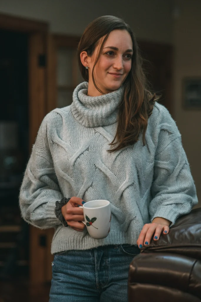Smiling woman in a pale grey-blue cable-knit turtleneck sweater and blue jeans holding a mug with a holly print