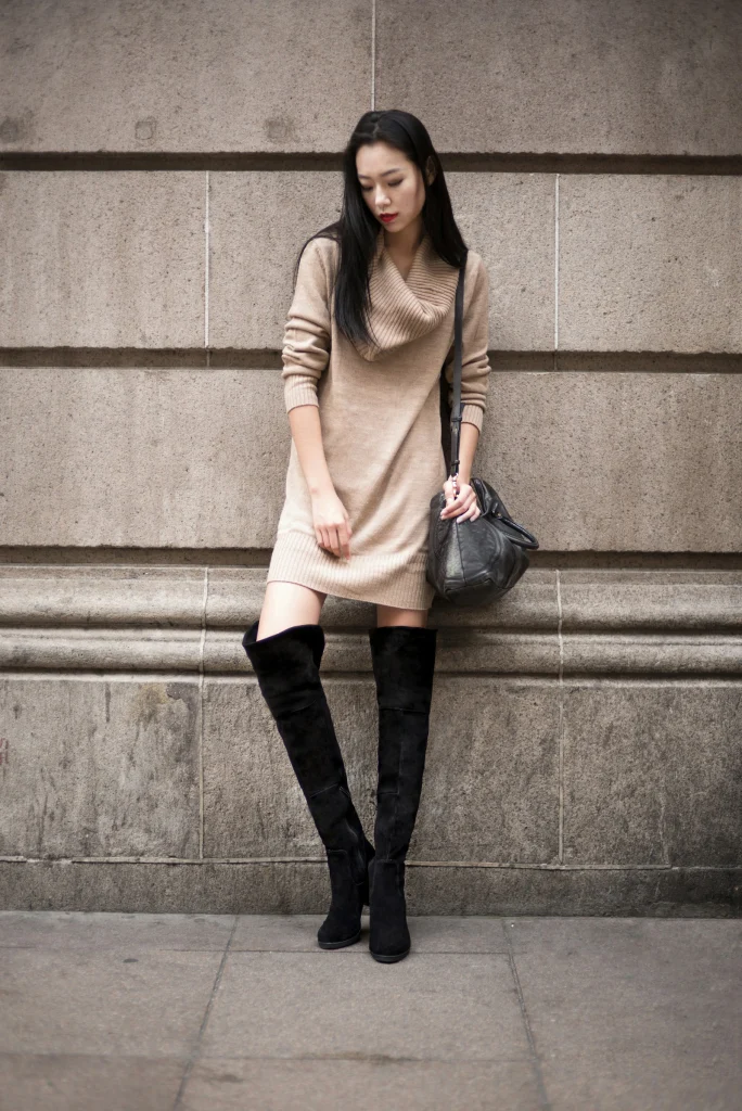 Woman in a camel cowl-neck sweater dress, black suede over-the-knee boots and a black slouchy shoulder bag leaning against a stone wall