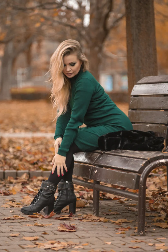 Woman in a dark green ribbed knit dress, black tights and black lace-up heeled ankle boots sitting on a park bench among autumn leaves