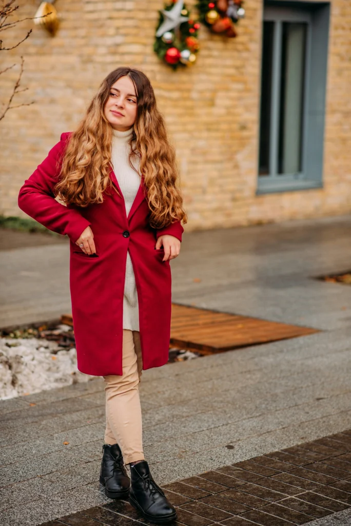 Woman in a red wool coat, long cream turtleneck knit, beige trousers and black lace-up boots walking past a wall decorated with Christmas wreaths