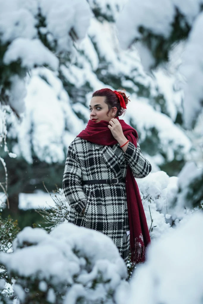 Woman in a belted black and white check wool coat and a long burgundy scarf standing among snow-covered trees