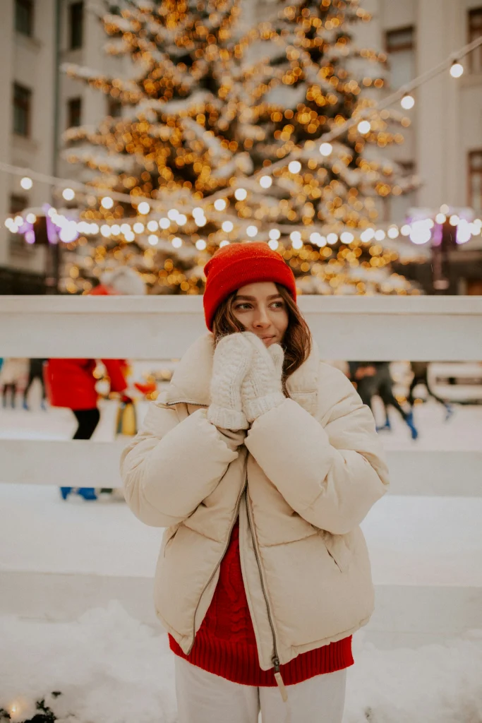 Woman in a cream puffer jacket over a red knit sweater with a red beanie and white knit mittens by an outdoor skating rink with a lit Christmas tree