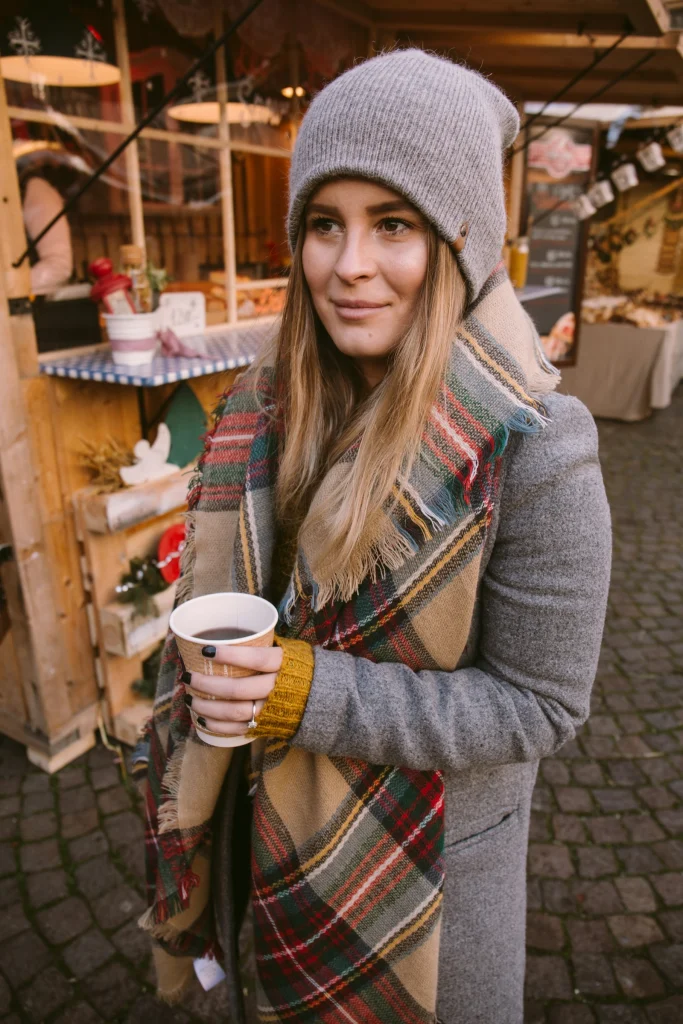 Woman at an outdoor Christmas market in a grey wool coat, tartan blanket scarf and grey beanie, holding a hot drink