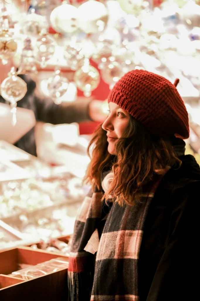 Woman in a red knit beret and black and red check scarf looking at glass ornaments at a Christmas market stall