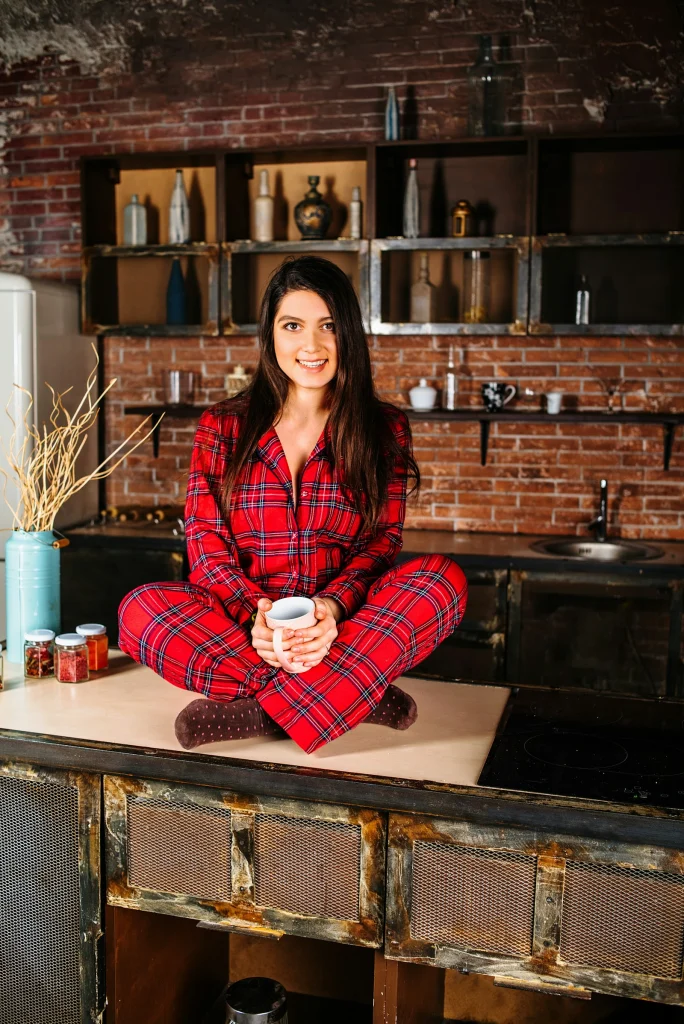 Woman sitting cross-legged on a kitchen counter in a red tartan flannel pajama set and socks, holding a mug