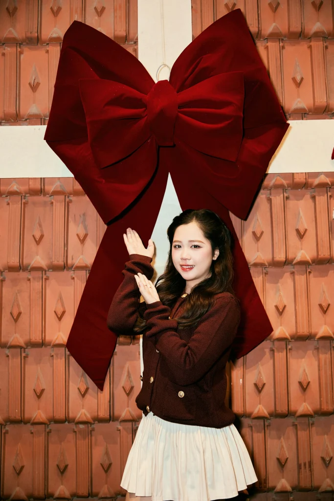 Woman in a burgundy cardigan with gold buttons and a cream pleated mini skirt posing under a giant red velvet bow