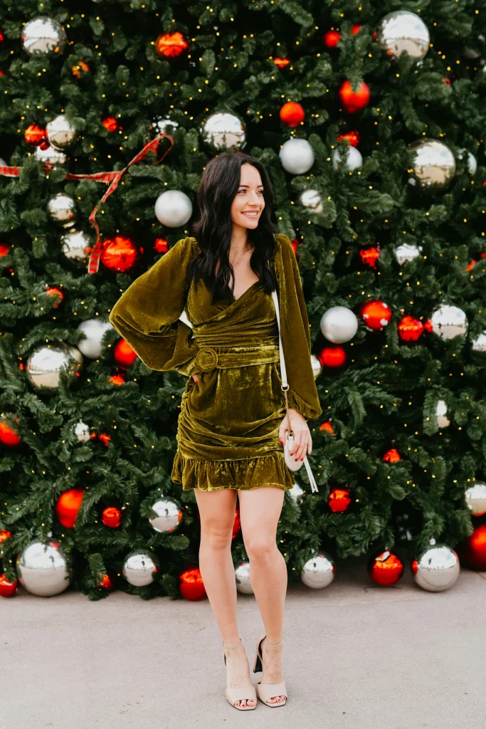 Smiling woman in an olive velvet wrap mini dress with nude block-heel sandals and a small white crossbody bag in front of a decorated Christmas tree