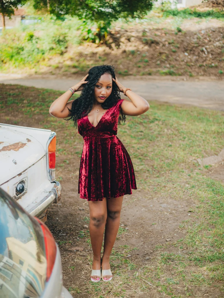 Woman in a red crushed-velvet skater dress and clear-strap sandals standing on grass beside a vintage car