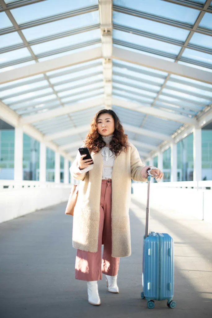 Woman with a blue carry-on suitcase in a cream shearling-trim coat, white blouse, dusty pink wide-leg trousers and white ankle boots walking through a glass walkway